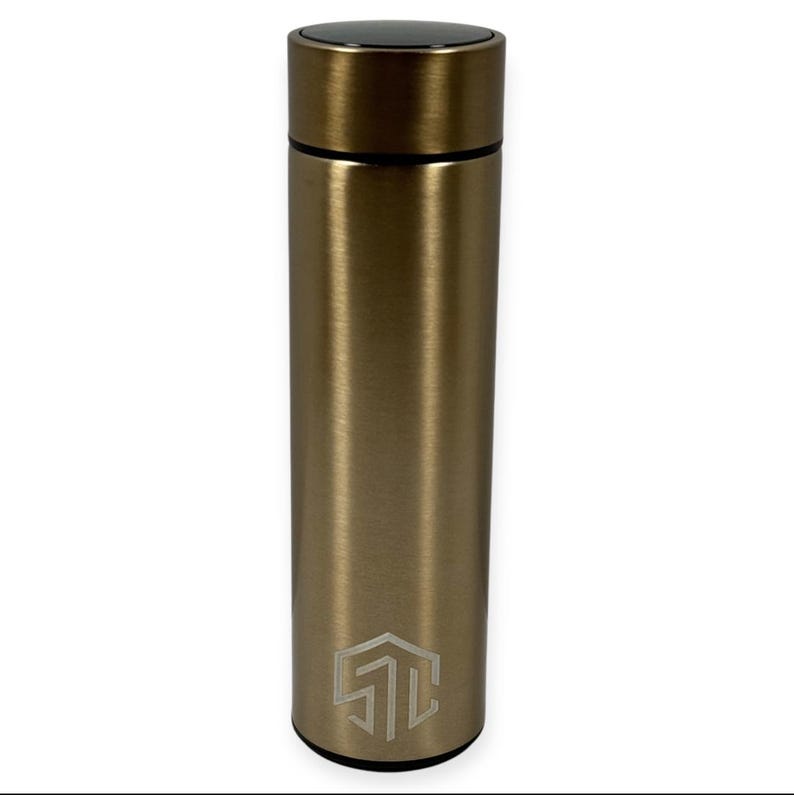 May include: A gold-colored, cylindrical water bottle with a black cap. The bottle has a brushed metal finish and a geometric logo near the base. The bottle is designed to keep beverages cold or hot.