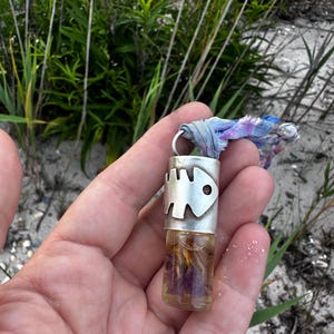 Könnte beinhalten: Ein kleines, klares Glasfläschchen gefüllt mit getrockneten Blumen und Flüssigkeit, gekrönt von einer silbernen Kappe mit Fischgrätenmuster. Ein Stoffband ist an der Kappe befestigt. Das Fläschchen wird in einer Hand vor dem Hintergrund von Sand und Grün gehalten.