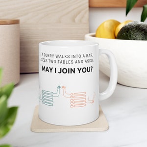 May include: White ceramic mug with the text "A QUERY WALKS INTO A BAR, SEES TWO TABLES AND ASKS: MAY I JOIN YOU?" in black. The mug features a humorous illustration of two tables with arms and hands.