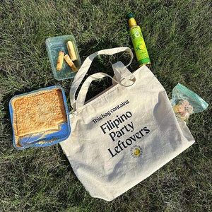 Può includere: Una borsa tote bianca con testo nero che recita "This bag contains Filipino Party Leftovers". La borsa è appoggiata sull'erba verde con una bottiglia di vetro trasparente, un contenitore di cibo e un sacchetto di plastica con del cibo dentro.