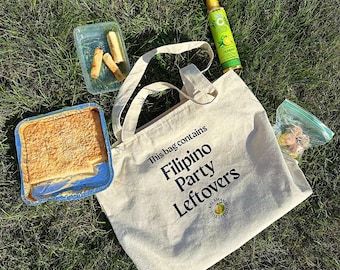May include: A white tote bag with black text that reads "This bag contains Filipino Party Leftovers". The bag is sitting on green grass with a clear glass bottle, a container of food, and a plastic bag with food inside.