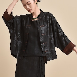 Könnte beinhalten: Eine schwarze Jacke mit dezentem Blumenmuster, braunen Ärmelbündchen und Knebelverschlüssen, kombiniert mit einem passenden schwarzen ärmellosen Oberteil und einem langen, geraden schwarzen Rock mit seitlicher Bindung und Schlitz, dazu schwarze Slipper.