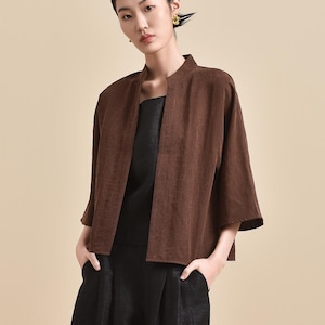 May include: A woman wearing a brown, short-sleeved jacket over a black top and wide-leg black pants. The outfit is a minimalist style, with a neutral background.