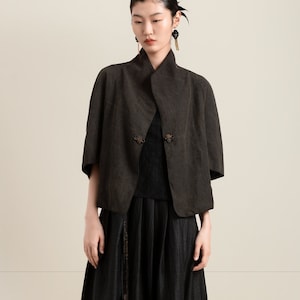 May include: A woman wearing a dark brown linen jacket with a black skirt and black leather shoes. The jacket has a mandarin collar and decorative buttons.