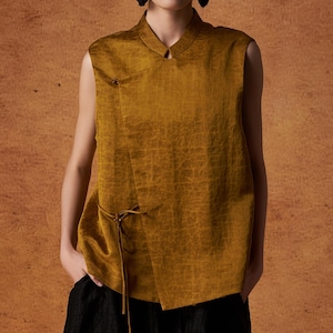 Könnte beinhalten: Senfgelbes ärmelloses Oberteil mit Stehkragen und Wickeloptik. Das Oberteil hat einen kleinen Knopfverschluss und ein Bindedetail an der Taille. Kombiniert mit einer schwarzen Hose mit weitem Bein hat das Outfit eine minimalistische Ästhetik.
