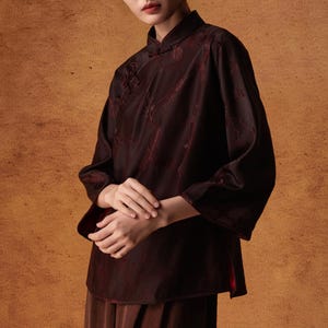 May include: A dark burgundy blouse with a mandarin collar and three-quarter sleeves. The blouse features a subtle floral pattern and is paired with matching brown pants. The outfit is a traditional style, suitable for formal or semi-formal occasions.