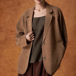 Puede incluir: Una mujer con una blazer marrón sobre una blusa verde oscuro y pantalones marrones. La blazer tiene un patrón texturizado y dos botones. El fondo es marrón texturizado.