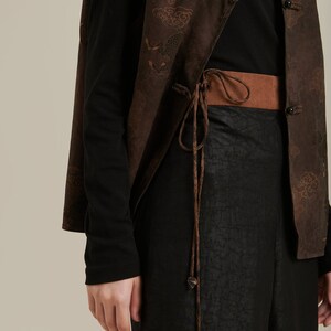Puede incluir: Una camisa negra de manga larga con un chaleco estampado marrón atado a la cintura con un cordón marrón. El chaleco tiene un diseño de dragón y nubes.