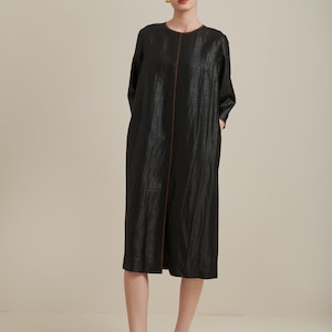 May include: A black midi dress with a brown trim and a subtle sheen. The dress has a round neckline and three-quarter sleeves. The model is wearing black leather slip-on shoes.