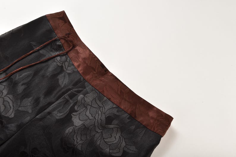 Op de afbeelding: Zwarte broek met een bruin bloemenpatroon en een bruine satijnen tailleband.