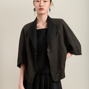 May include: A woman wearing a black linen jacket with a button closure and a black skirt. She is wearing long black earrings with gold accents.