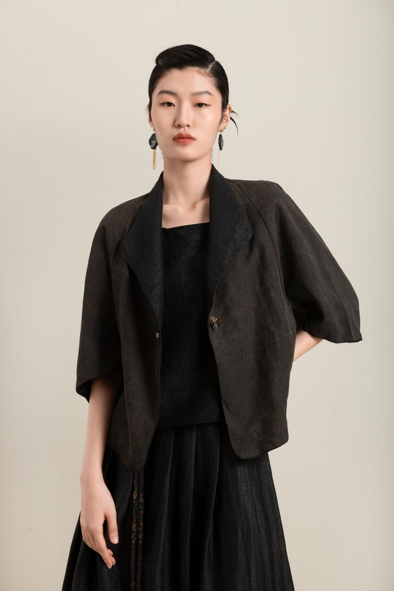 May include: A woman wearing a black linen jacket with a button closure and a black skirt. She is wearing long black earrings with gold accents.