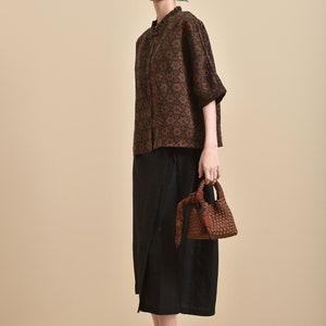 May include: A woman wearing a brown and black patterned blouse with a black skirt and black shoes. She is carrying a brown woven bag with a black scarf.
