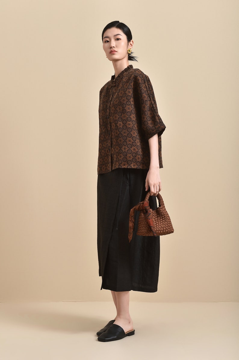 May include: A woman wearing a brown and black patterned blouse with a black skirt and black shoes. She is carrying a brown woven bag with a black scarf.