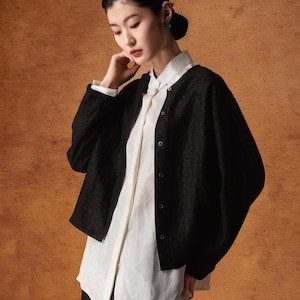 Puede incluir: Una mujer con una chaqueta negra texturizada con botones sobre una camisa blanca con botones. La chaqueta tiene mangas largas y cuello redondo. La mujer también lleva pantalones negros. El fondo es de color marrón cálido.