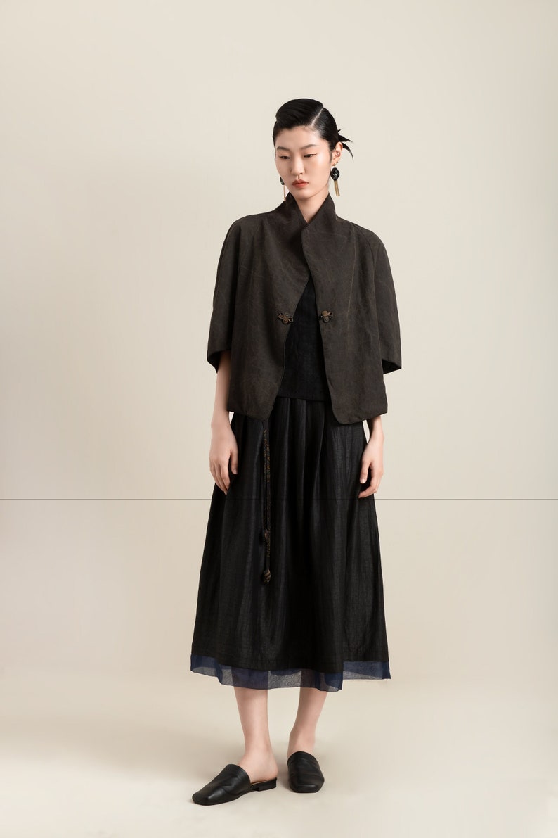 May include: A woman wearing a dark brown linen jacket with a black skirt and black leather shoes. The jacket has a mandarin collar and decorative buttons.
