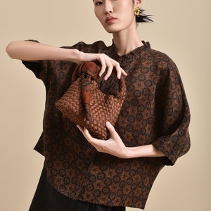 May include: A woman wearing a brown and black patterned shirt with a brown woven bag. The bag has a brown and orange patterned scarf tied around the handle.