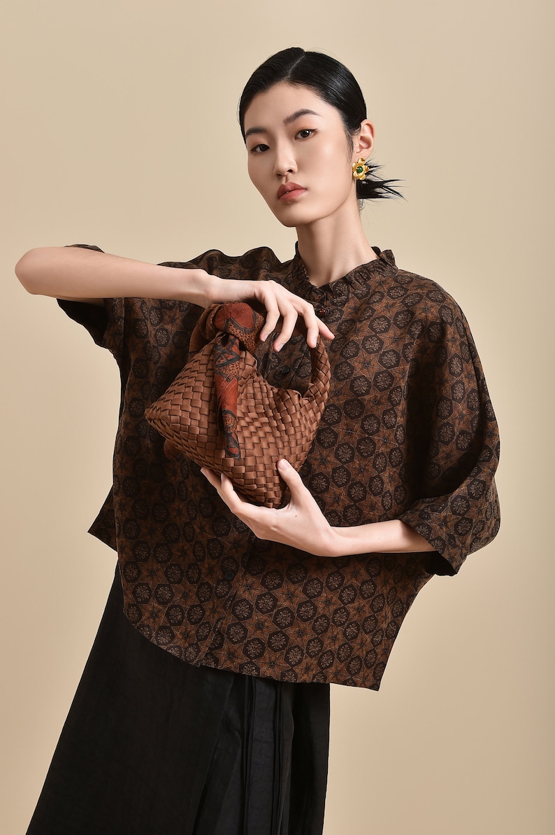 May include: A woman wearing a brown and black patterned shirt with a brown woven bag. The bag has a brown and orange patterned scarf tied around the handle.