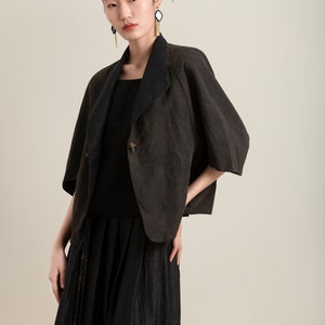 May include: A woman wearing a black top and a brown linen jacket with a black collar. The jacket has a single button closure and a slightly flared silhouette. She is also wearing a long black skirt with a tassel detail.