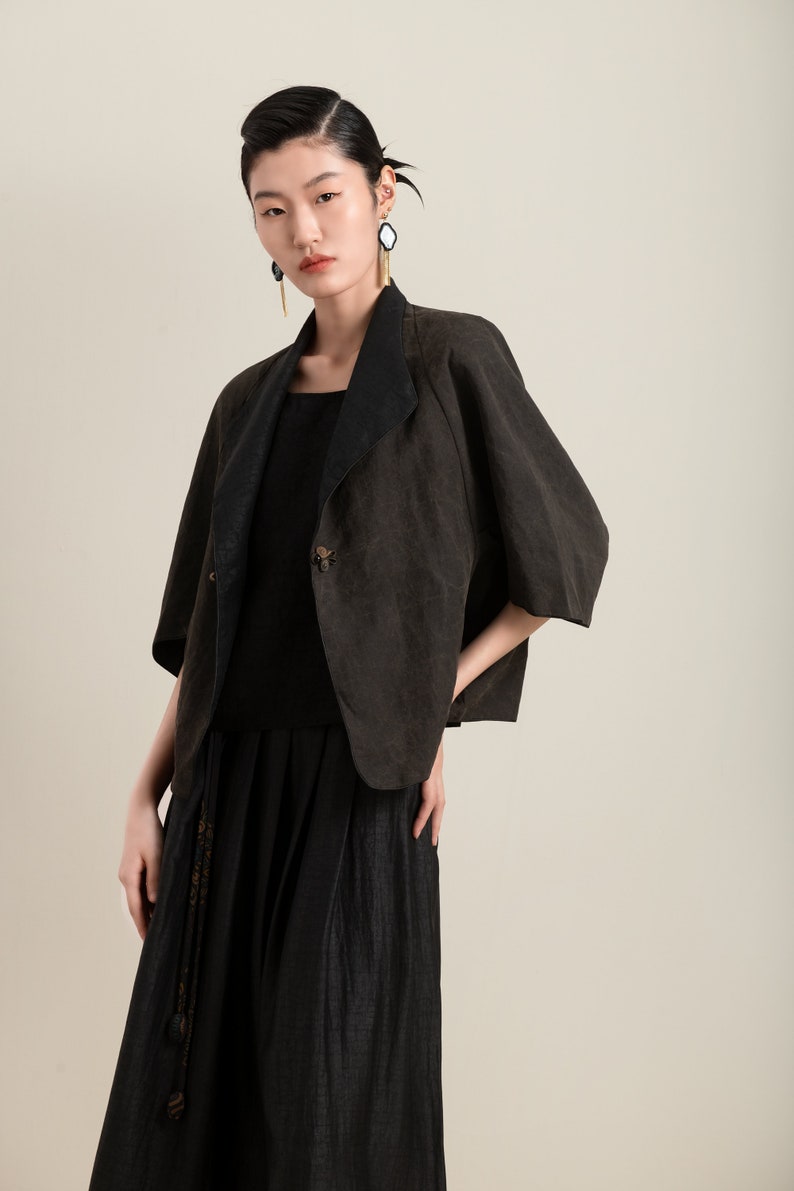 May include: A woman wearing a black top and a brown linen jacket with a black collar. The jacket has a single button closure and a slightly flared silhouette. She is also wearing a long black skirt with a tassel detail.