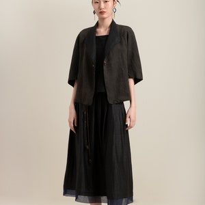 May include: A woman wearing a black linen jacket with a black skirt and black shoes. The jacket has a collar and a button closure. The skirt is a midi length and has a slight A-line shape. The shoes are flats with a pointed toe.