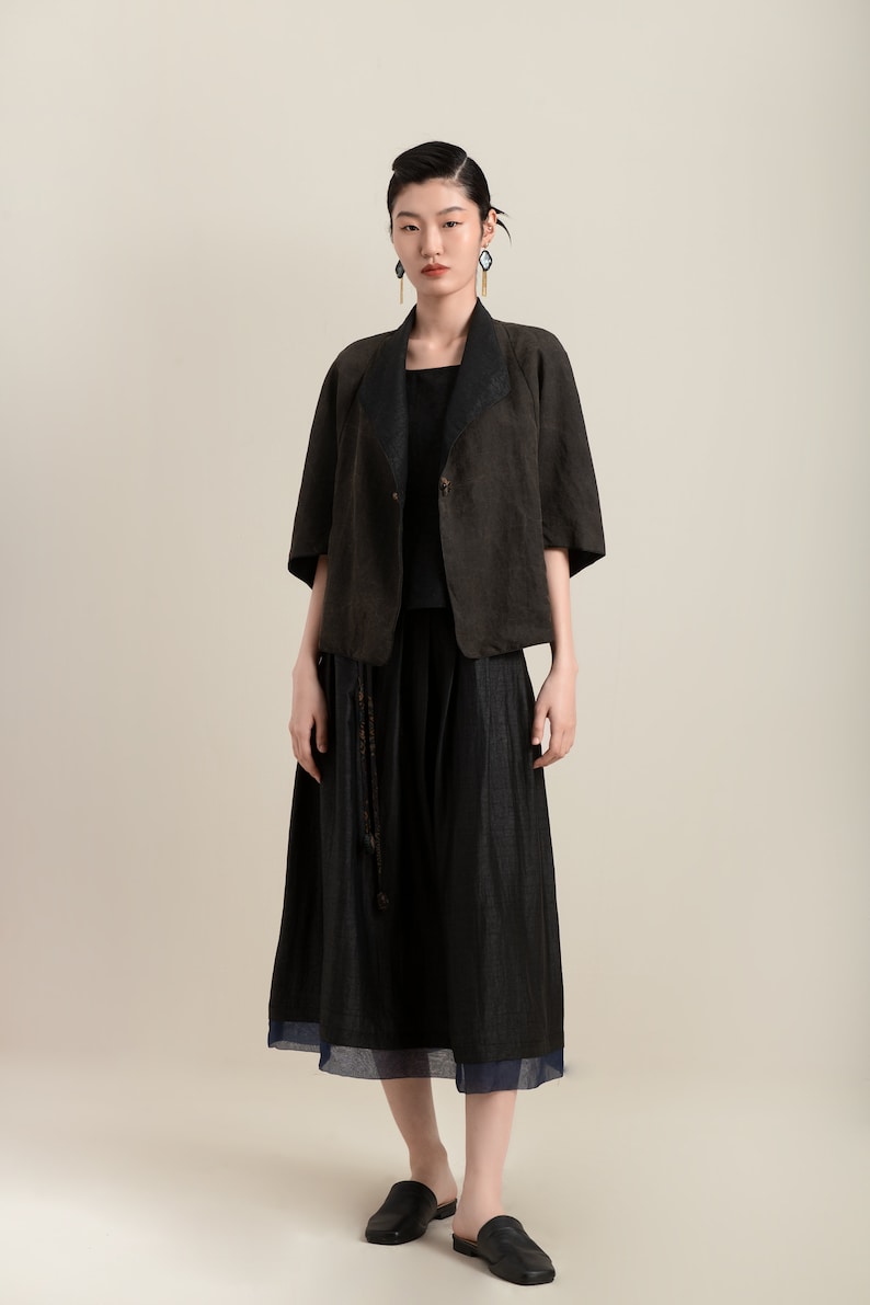 May include: A woman wearing a black linen jacket with a black skirt and black shoes. The jacket has a collar and a button closure. The skirt is a midi length and has a slight A-line shape. The shoes are flats with a pointed toe.