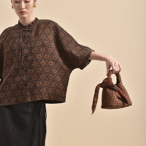 May include: A woman wearing a brown and black patterned blouse with a black skirt and a brown woven bag with a brown and orange patterned scarf tied to the handle.