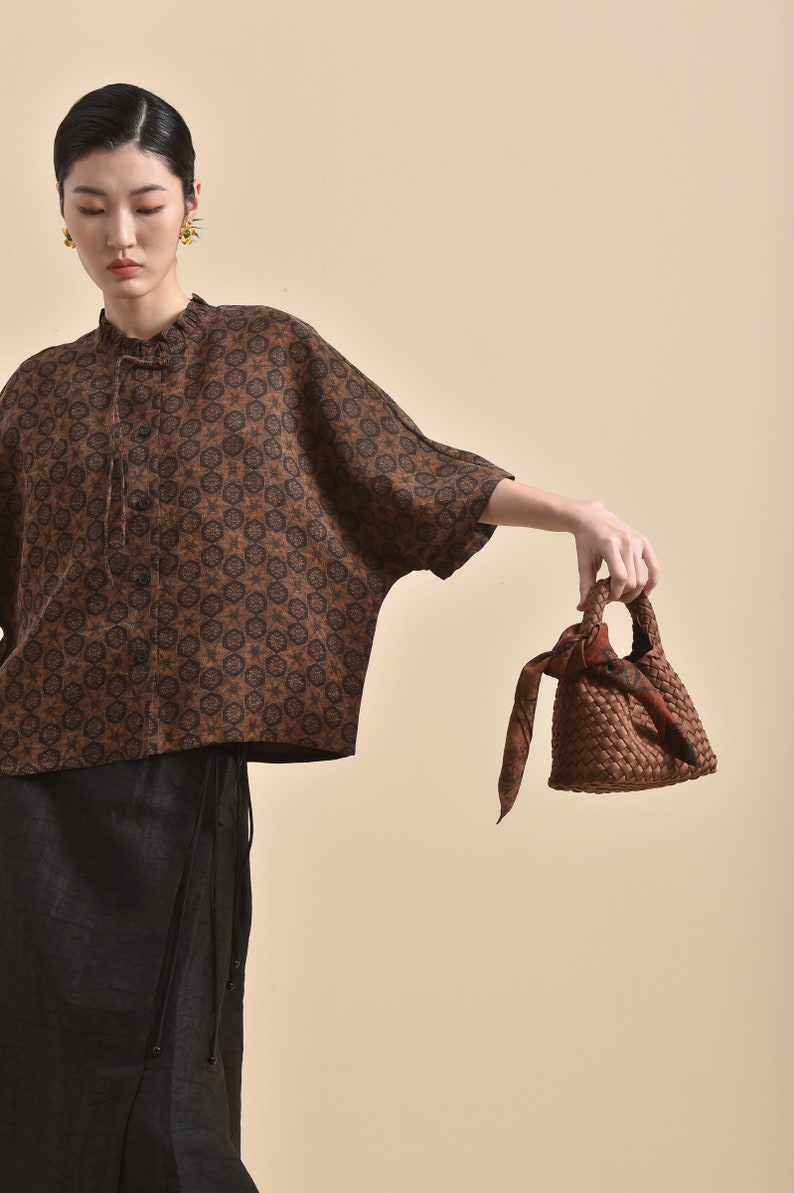 May include: A woman wearing a brown and black patterned blouse with a black skirt and a brown woven bag with a brown and orange patterned scarf tied to the handle.