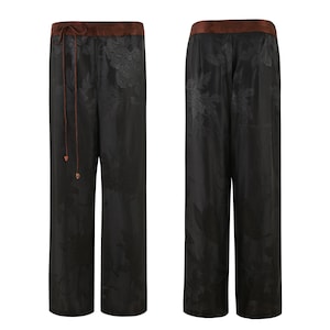 Op de afbeelding: Zwarte wijde broek met een bloemenpatroon en een bruine tailleband. De broek heeft een trekkoord.