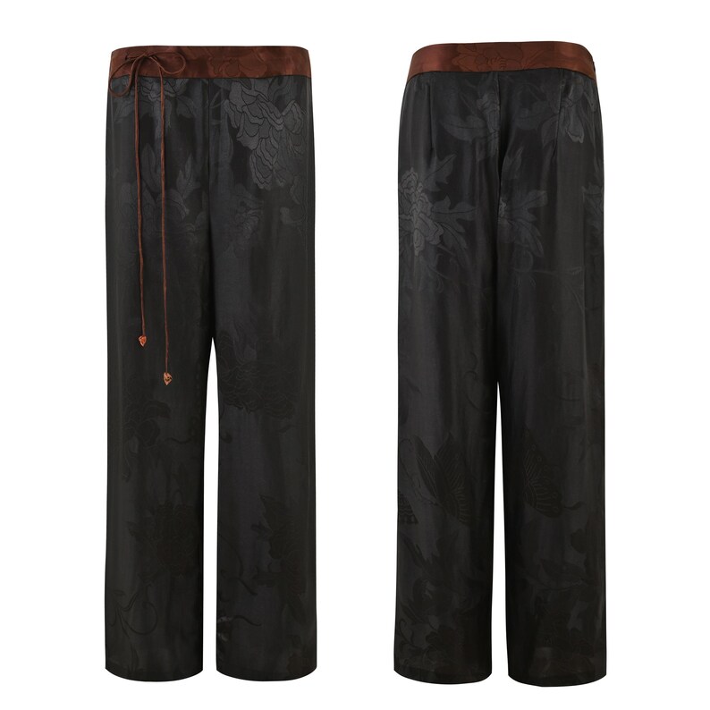 Op de afbeelding: Zwarte wijde broek met een bloemenpatroon en een bruine tailleband. De broek heeft een trekkoord.
