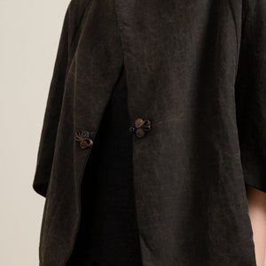 May include: A black, short-sleeved jacket with a unique, asymmetrical design. The jacket has a collar that folds over and a decorative button closure. The fabric has a textured, crinkled look.