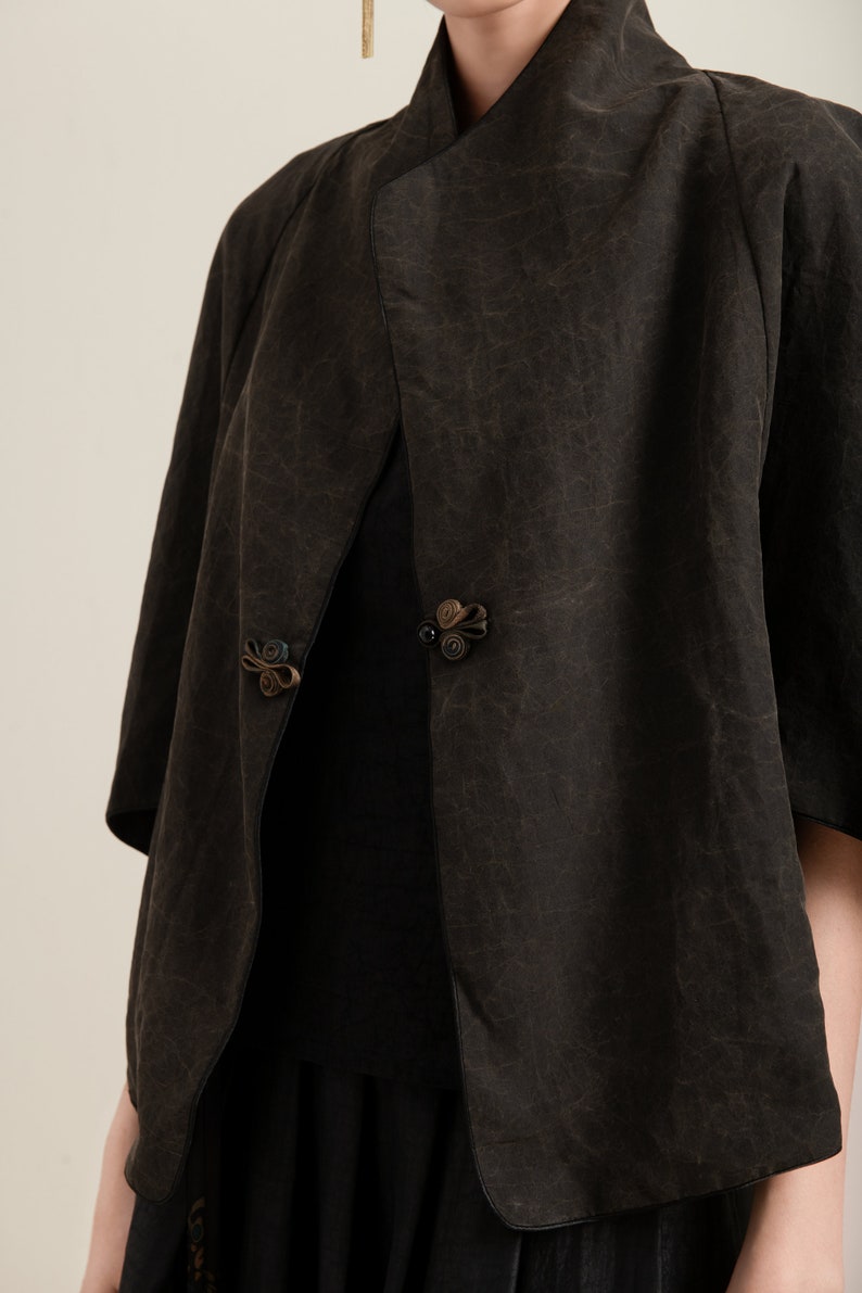 May include: A black, short-sleeved jacket with a unique, asymmetrical design. The jacket has a collar that folds over and a decorative button closure. The fabric has a textured, crinkled look.