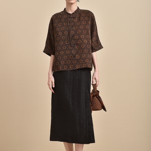 May include: A brown and black patterned short-sleeved shirt with a mandarin collar and a black skirt. The model is wearing black slip-on shoes and carrying a brown woven bag.