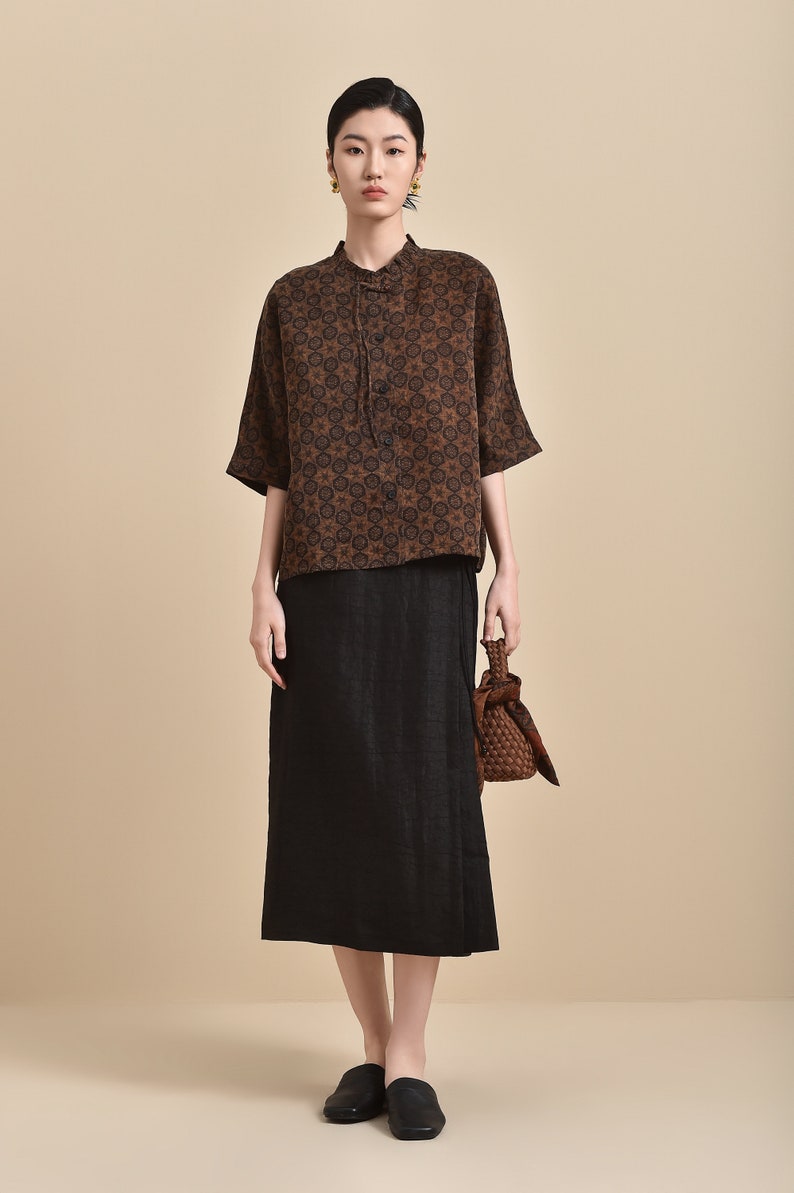 May include: A brown and black patterned short-sleeved shirt with a mandarin collar and a black skirt. The model is wearing black slip-on shoes and carrying a brown woven bag.