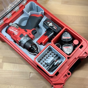 Puede incluir: Una caja de herramientas roja y gris con un taladro Milwaukee, una batería y una variedad de brocas. La caja tiene una tapa transparente y un asa.