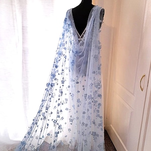 May include: A light blue bridal cape with a floral pattern. The cape is made of sheer fabric and drapes over a mannequin. The cape features a deep V-neck and a flowing design, suitable for a wedding or special event.