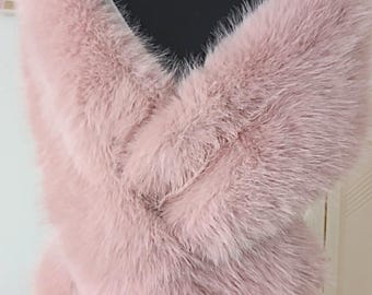 May include: A soft, dusty pink faux fur stole draped over a black mannequin. The wrap features a thick, plush texture with long, dense fibers that create a voluminous appearance, covering the shoulders and crossing over the chest area.