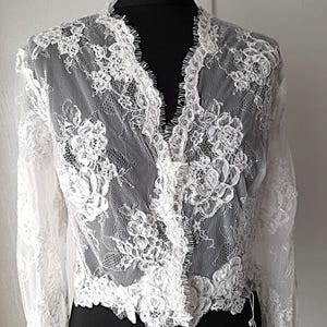 Puede incluir: Un top de encaje blanco con mangas largas y cuello en V. El delicado estampado floral cubre toda la prenda. El top se exhibe en un maniquí negro, mostrando los intrincados detalles del encaje.