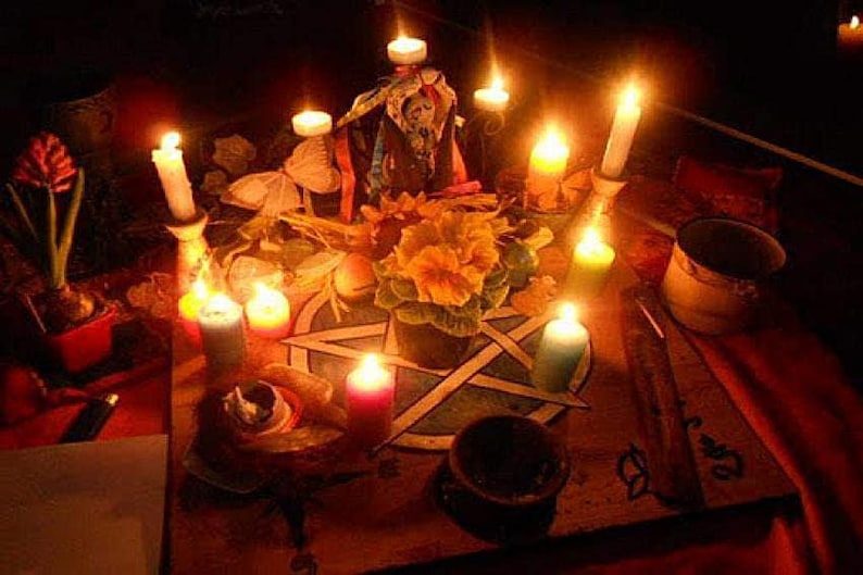 May include: A dimly lit scene featuring a table adorned with candles, flowers, and a pentagram. Various candles of different colors are lit, casting a warm glow. A small pot of flowers and other ritualistic items are present.