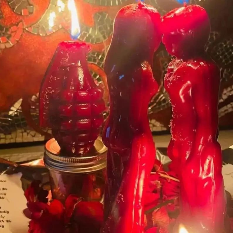 May include: A collection of red candles, including a grenade-shaped candle and two figure-shaped candles. The candles are lit, with flames visible. The background includes a mosaic design and a jar.