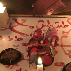 May include: Two handmade dolls with painted faces and fabric clothing, connected by a red band. The dolls are set against a wall with red painted Arabic script. A lit candle and a dark, round object are in the foreground.