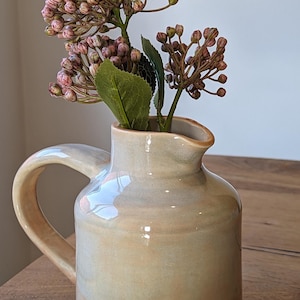 以下が含まれることがあります： 丸みを帯びたボディとハンドルが付いた薄いベージュのセラミックピッチャー。ピッチャーにはピンクの花束が入れられています。