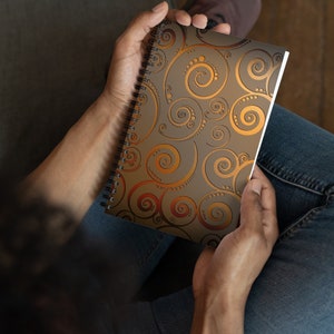 Könnte beinhalten: Ein spiralgebundenes Notizbuch mit einem braunen und goldenen Wirbelmuster auf dem Einband.