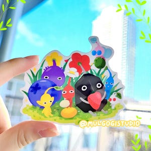 Op de afbeelding: Een sticker met een groep kleurrijke cartoonfiguren, waaronder een paars wezen, een rood wezen, een zwart wezen dat een aardbei vasthoudt en een geel wezen. De figuren zijn omringd door bloemen en bladeren. De sticker is op een witte achtergrond en heeft de tekst "@mulgogistudio" in de rechterbenedenhoek.