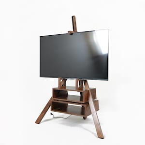 May include: A wooden TV stand with a large, black screen. The stand has an easel design with angled legs and built-in shelves. The wood has a warm, brown tone, and the stand is set against a white background. The stand is approximately 150 cm tall.