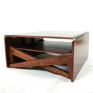 May include: A square wooden coffee table with a dark brown finish, featuring a flat top surface, a lower storage shelf, and side panels with a distinct crisscross geometric cutout design that connects the base to the tabletop structure.