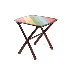 May include: A folding table with a dark brown wooden frame and a square tabletop featuring diagonal slats in a rainbow color palette. The tabletop surface displays stripes of red, orange, yellow, green, blue, and purple, supported by a cross-leg base.