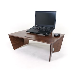May include: A dark brown wooden laptop desk with a rectangular top and angled side supports. The desk features a cutout handle on the surface and holds a black laptop computer and a computer mouse on its flat, smooth, stained wood surface.
