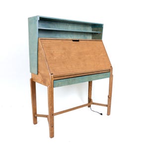 May include: A wooden bureau desk featuring a natural brown finish on the main body and a blue-stained finish on the upper hutch and drawer front. The desk includes a fold-down writing surface, an open shelf, and four legs with crossbars.
