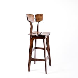 May include: A tall bar stool crafted from dark brown wood, featuring four straight legs with connecting support bars. The seat is divided into two distinct sections, and the backrest consists of two separate, rounded rectangular panels.